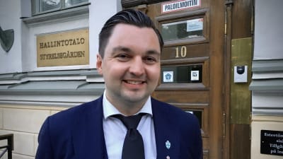 Man i blå kavaj står utanför en gul byggnad.