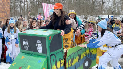 En pulka är ombyggd till en grön lastbil, och åker ner med glada studerande på en snöig gata. Många andra ungdomar står bredvid och hejar.