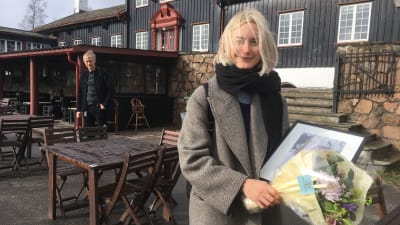 Vaaleahiuksinen nuori nainen, jonka hiuksia tuuli pöllyttää, seisoo puisen rakennuksen edessä ja taka-alalla mies katsoo maahan