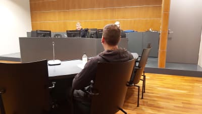 En ung man sitter på en stol i en rättssal, ryggen mot betraktaren. Han står åtalad för grovt skattebegrägeri i vad som är Finlands första Onecoin-rättegång.