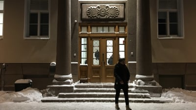 Entre till stor stenbyggnad. Trappor och utsmyckad dörr. Vid trappan snö och person som vandrar förbi.