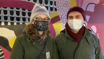 Emilia och Timo står med munskydd framför en färgglad vägg