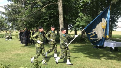 Tre militärklädda män håller en blå fana med bilden av ett vitt Gotlandsfår.