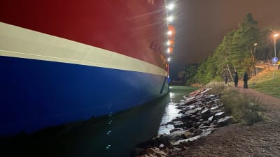 Viking Grace som kört på grund nära Mariehamn. Man ser ur nära stranden färjan är. Vid strandpromenaden går tre personer.