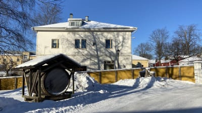En gammal tunna står utställd på en innergård. Området är omgärdat med staket. I bakgrunden syns en äldre byggnad.