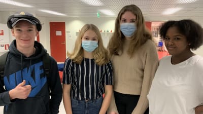 Fyra unga personer står intill varandra i skolmiljö, tittar in i kameran och ler. Två av dem bär munskydd.