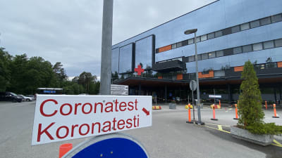 En skylt till corona provtagning. I bakgrunden ses en sjukhusbyggnad.