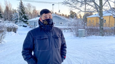 En man i vinterjacka och svart munskydd står på en snöig gård. I bakgrunden syns en gata och några småhus.