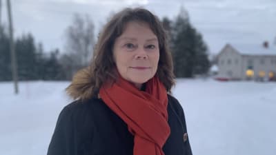 Kvinna med ytterrock och roströd halsduk står ute i vintrigt landskap. Tittar leende in i kameran.