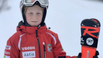 En pojke med röd skiddräckt, hjälm och skidglasögon står med ett par skidor i hand