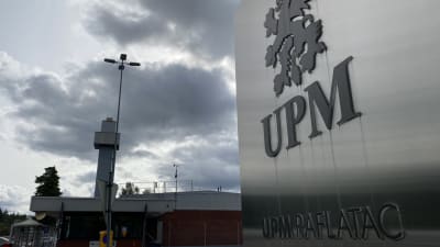 UMP Raflatacin metallinen kyltti, jonka takaa näkyy tuotantolaitos ja harmaa taivas