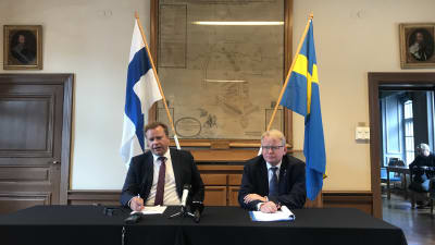 Antti Kaikkonen och Peter Hultqvist sitter vid ett bord med Finlands och Sveriges flaggor i bakgrunden.