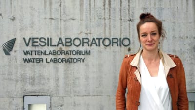 Julia Talvitie står vid en betongvägg där det står "Vesilaboratorio, vattenlaboratorium". Julia har brun läderjacka och en vit skjorta på sig.