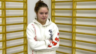En cirka fjortonårig licka står med armarna i kors i en gymnastiksal. Hon har en vit hoodie med text i rött. Bakom träribbor som man kan klättra i.