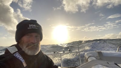 Yksinpurjehtija Ari Huusela veneessään kaukana merellä Vendée Globe -kilpailussa. Taustalla iso aallokko ja aurinko paistaa. 
