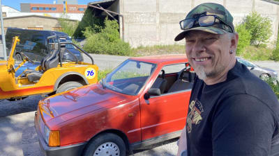 Man med skärmmössa framför liten röd bil.