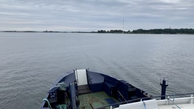 Fotot är taget på ett fartyg, på väg mot Mjölö. En del av den blåa fören på båten syns, och i bakgrunden ser man klippor som tillhör Mjölö. Det är annars mulet och grått.