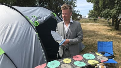 En man i kostym med strykjärn i handen utanför ett tält.