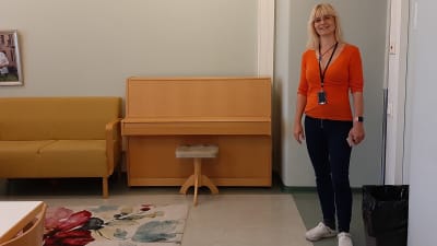 Blond långhårig kvinna i glasögon, orange blus och svarta byxor står vid ingången i ett rum med piano.