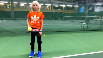 En ung pojke i orange sjokrta håller i en tennisracket och boll.