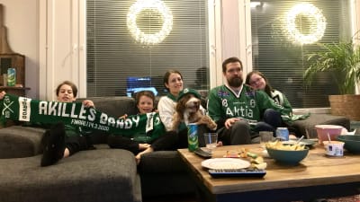 En familj med mamma, pappa och tre barn sitter i en soffa med en hund. De har gröna mössor och halsdukar på sig.
