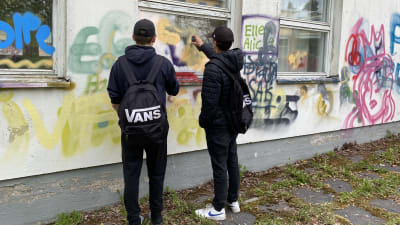 Två unga killar sprejar grafitti på grå skolbyggnad.