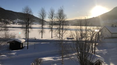 Vinterlandskap i Norge. Sol, snö och små hus nära isbelagd sjö. I bakgrunden fjäll.