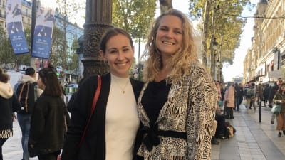 Två kvinnor står på en gata i Paris. De håller varandra om ryggen och ler. 
