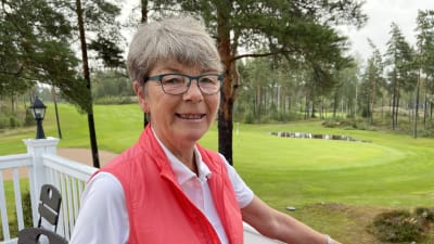 Porträtt av Annika Colérus som står med armen på räcket till klubbhuset vid Eke golf. Hon ser in i kameran och ler. I bakgrunden skymtar golfplanen och ett av vattenhindren. 