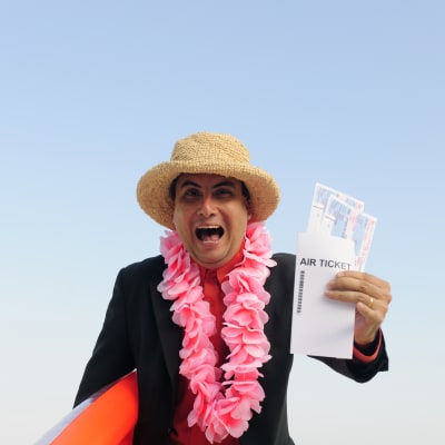 en medelålders kostymklädd man står på en strand med solhatt på och en flygbiljett i handen. Han ler ivrigt in i kameran.