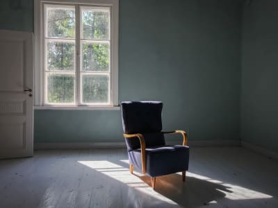 Sininen nojatuoli tyhjässä huoneessa, taustalla moniruutuinen vanha ikkuna, josta paistaa kesäinen aurinko.