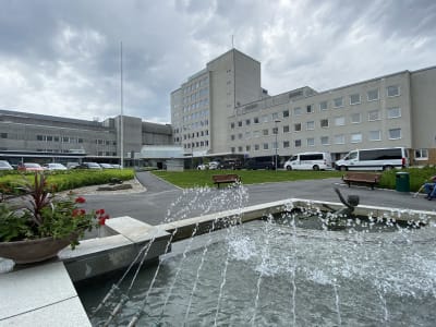 En sjukhusbyggnad. I förgrunden ses en fontän omgiven av ett grönområde.