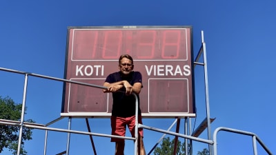En man med glasögon står vi en resultattavla uppe på en ställning. 