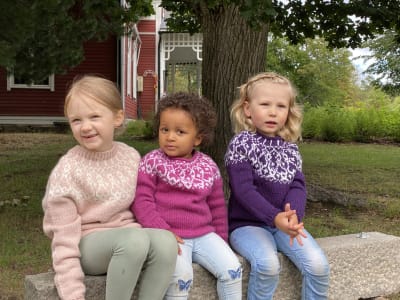 Tre flickor iklädda handstickade strömsöbarntröjor sitter på en bänk.