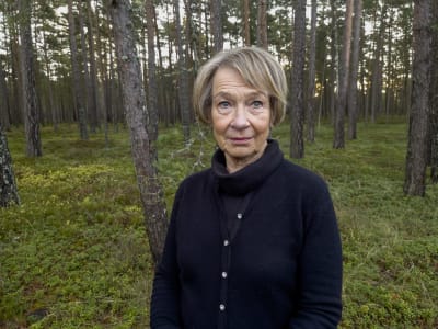 En kvinna i övre medelåldern står ute. Tallskog i bakgrunden.