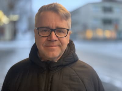 En man med grått hår och mörka bågar på glasögonen står i en urban miljö med vinterjacka. 