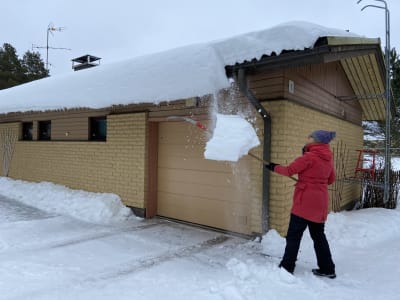 Nainen pudottaa lunta omakotitalon katolta. 