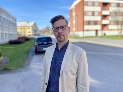 En man i beige kavaj och blå skjorta och glasögon står ute i solen.