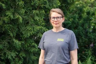 Kvinna som står framför ett buskage. Hon är iklädd glasögon och en grå t-skjorta med texten "Korkeasaari Zoo"