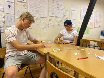 Två ungdomar sitter vid ett bord och skriver någonting på lappar. 