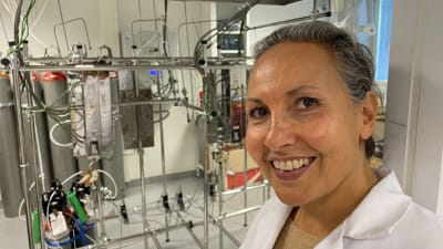 En leende kvinna står i ett laboratorium vid en massa slangar och ledningar. 