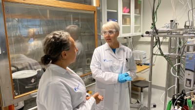 Två kvinnor med labb-rockar står i ett laboratorium intill en stor apparat med rör. 