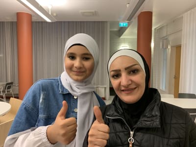 En flicka och en kvinna som båda ler och visar tummen upp. Båda bär hijab. I bakgrunden skolmatsal.