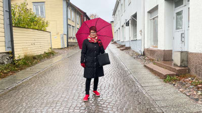 En kvinna med höstjacka och ett anilinrött paraply står på en kullerstensgata i Gamla Borgå.