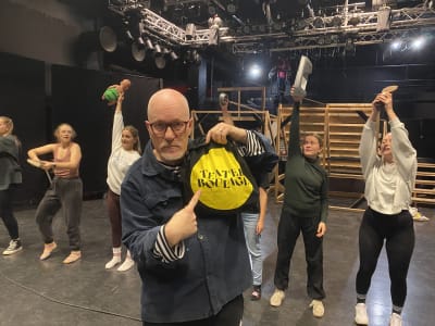 Skallig man med glasögon framför unga dansande kvinnor.
