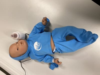 En baby-docka iklädd en blå tröja och blåa byxor. En sladd löper från dockan ur bilden. Bredvid dockan en nappflaska.