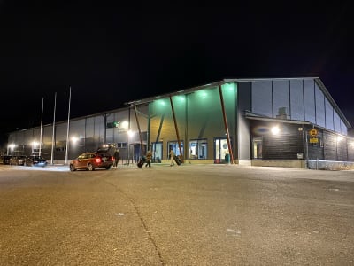 En grå hallbyggnad, kväll, byggnaden är upplyst. Bilar står parkerade framför byggnaden, barn på väg in i hallen. Raseborgs ishall.