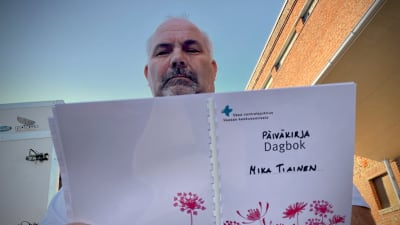Mika Tiainen läser i sin sjukhusdagbok. På pärmen står det "Dagbok" och så har någon för hand fyllt i "Päiväkirja" och "Mika Tiainen".