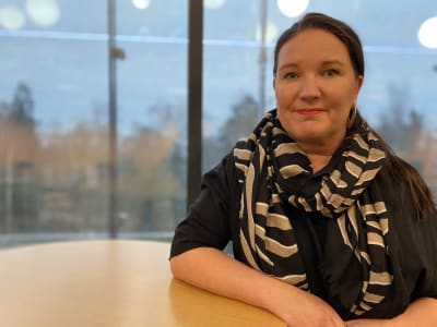 Kvinna sitter vid bord och tittar in i kameran.