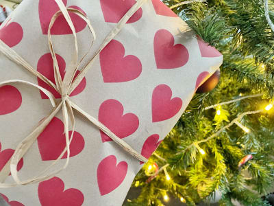 Ett paket packat i vitt papper med rosa hjärtan på lutar mot en julgran med julljus.